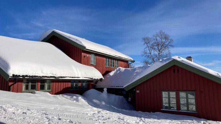 Rødt trehus med snødekt tak under blå himmel i norsk vinterlandskap.