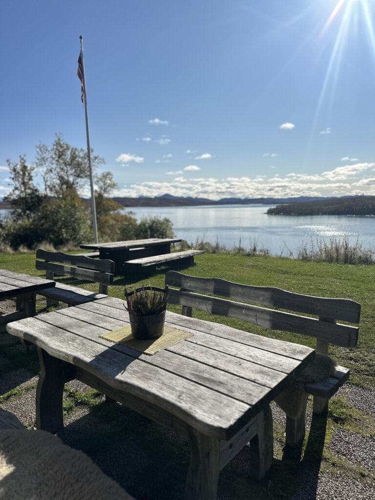 Utebord med gammeldags trebenk, norsk flagg og vakker innsjøutsikt på solfylt dag.