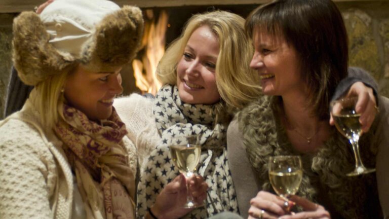 Tre kvinner nyter vin ved peisen i vinterklær og skjerf, med smil og vennskap.