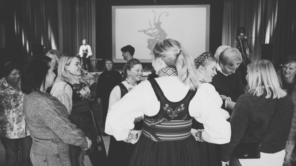 Gruppe mennesker i bunad på en samling, med tradisjonelle norske drakter. Skjerm i bakgrunnen.