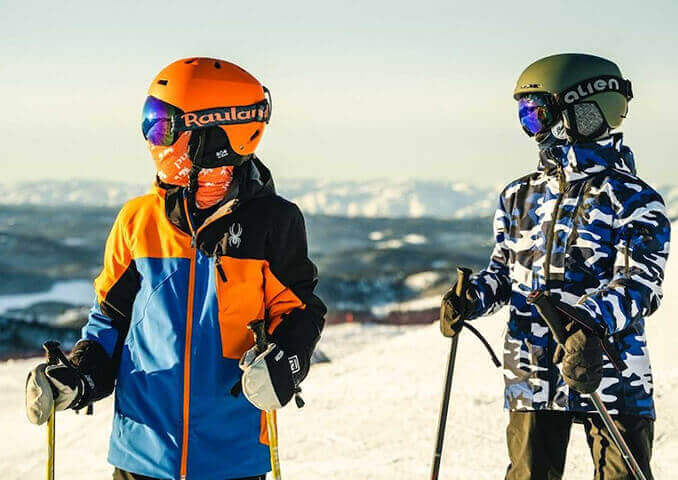 To barn på ski med hjelmer står på en snødekket fjelltopp i solfylt vær, klare for å stå på ski.