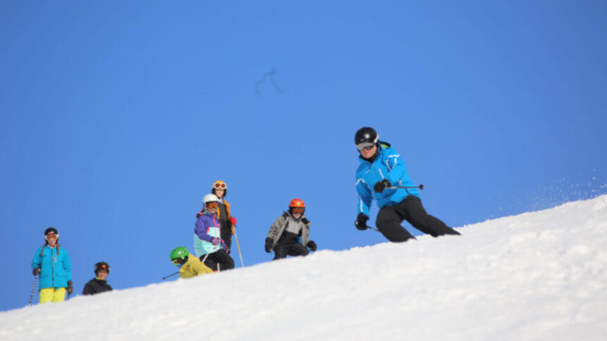 En gruppe mennesker står på en snødekket bakke og en person står på ski foran dem under en klar blå himmel.