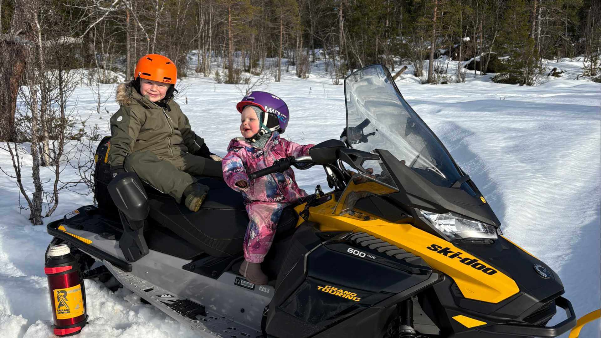 To barn smiler mens de sitter på en snøscooter i snødekt skog, klare for vintereventyr.