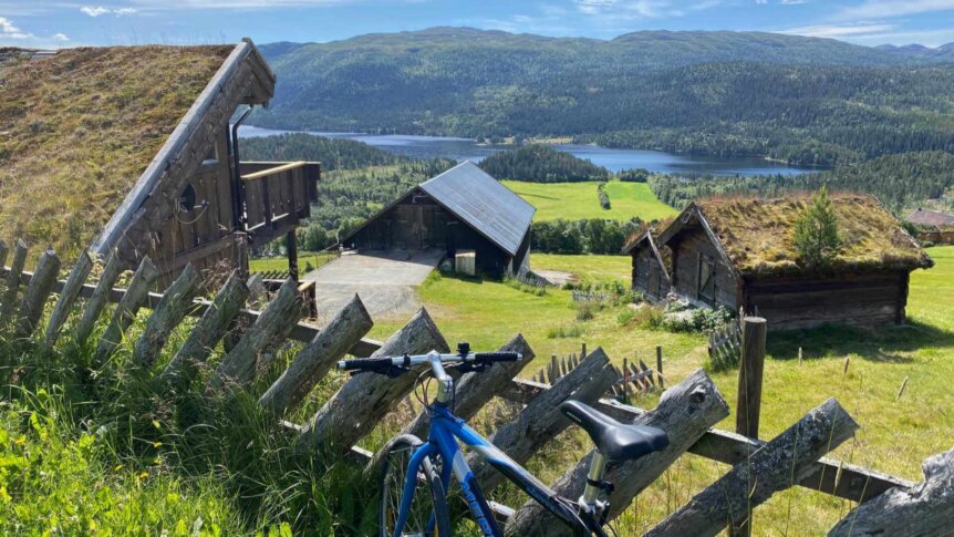 Bilde av en blå sykkel foran tradisjonelle hytter med grønt tak, omgitt av vakker norsk natur og fjellutsikt.