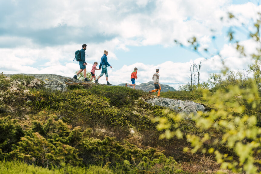 Familie på tur i naturen, bestående av voksne og barn, som går på en sti med grønt landskap rundt.