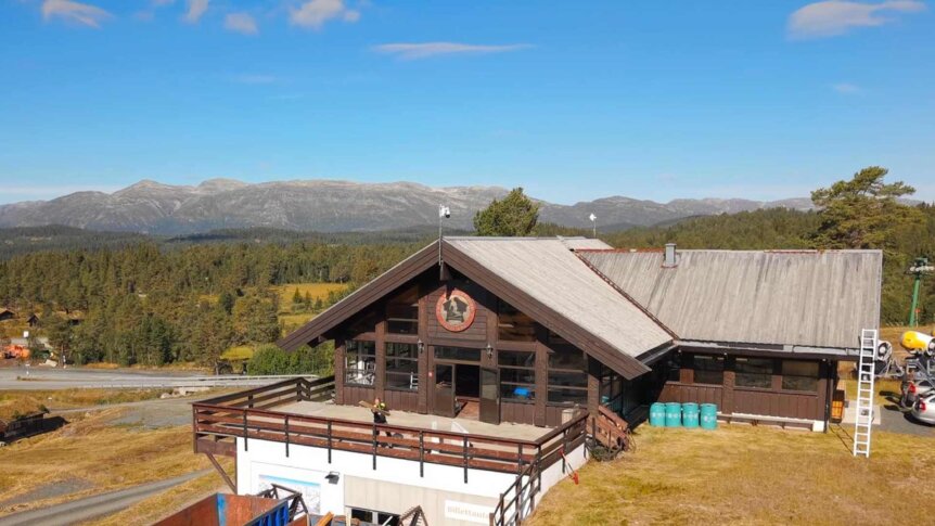 Alt-tekst: Utsikt over tradisjonell hytte med fjellbakgrunn og klar blå himmel i Norge.