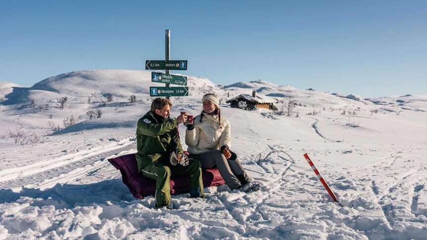 Par med varm drikke sitter på snødekt bakgrunn ved skilt for turgåing i fjellet. Klar himmel og fjelllandskap rundt.