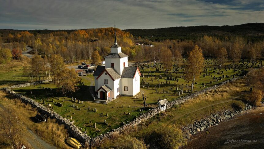 Historisk kirke omgitt av gravplasser og høstfargede trær i vakker natur. Perfekt for fotografering og sightseeing.