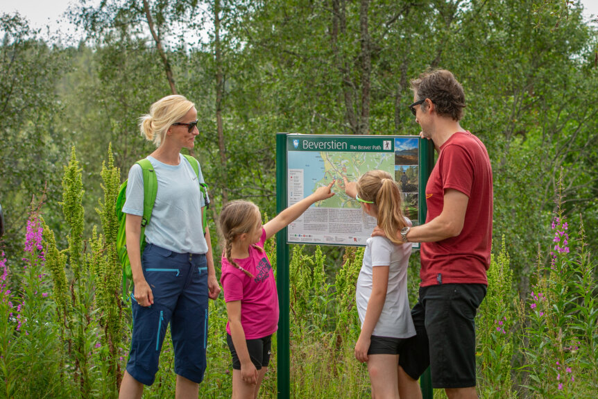 Familie ser på kart over Beverstien og peker på ruten, omgitt av frodig natur og blomster.