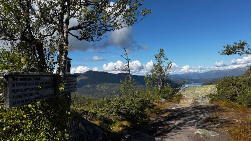 Utsikt fra dansersteinen med skilting om historien, omgivelsene og fjellene i bakgrunnen. Blå himmel og skyer.