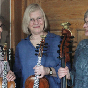 Tre kvinner holder instrumenter, smilende, foran en trevegg. Kulturelt uttrykk og glede for musikk.
