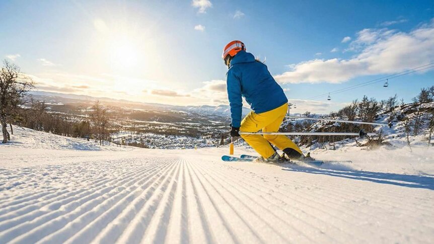 En person i blå jakke og gule bukser står på ski i en solfylt, snødekt bakke.