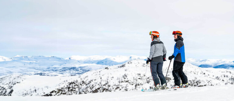 To mennesker på ski med utsikt over snødekte fjell, klare for en dag i bakken.