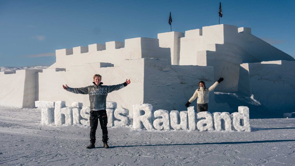 To personer poserer foran en stor snøslot med teksten This is Rauland i snøen. Vinterlandskapet er klart og solfylt.