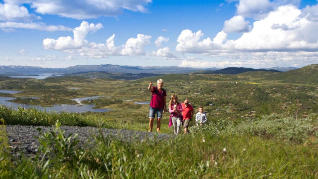 Familie på tur i norsk fjellandskap, med utsikt over innsjøer og grønne bakker under en blå himmel med skyer.
