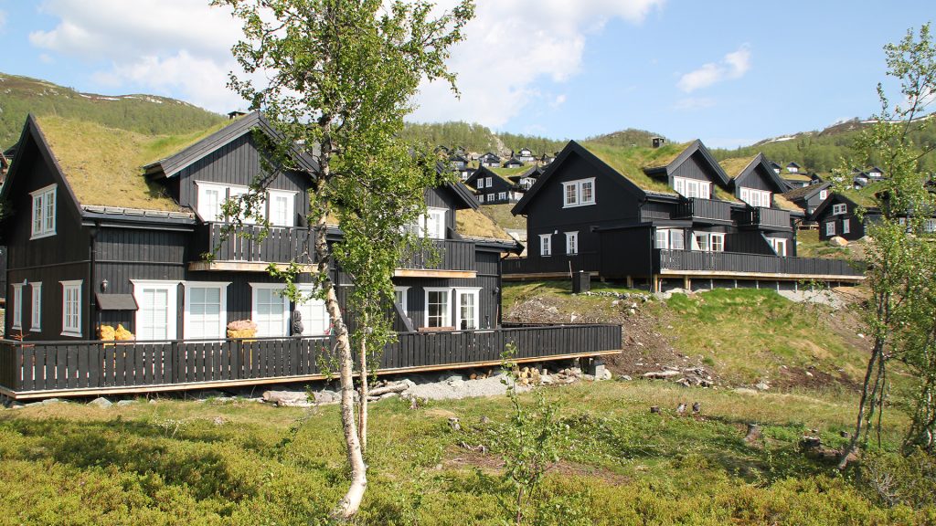 Svarte hytter i fjellandskap under blå himmel, omgitt av natur og fargerike trær, Norge.