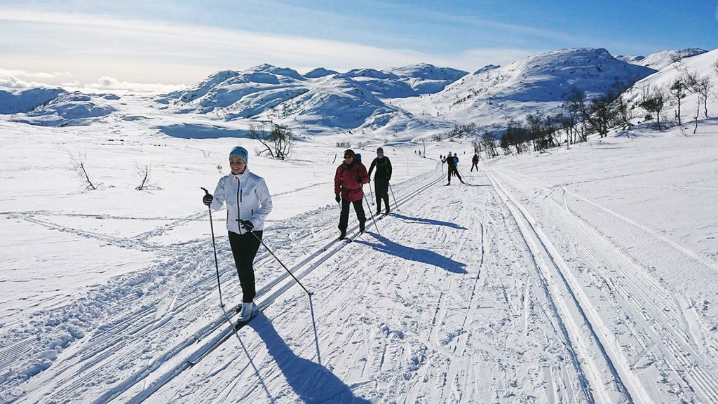 Mennesker går på langrenn i snødekte fjell. Klar blå himmel og solskinn gir perfekt skiføre.