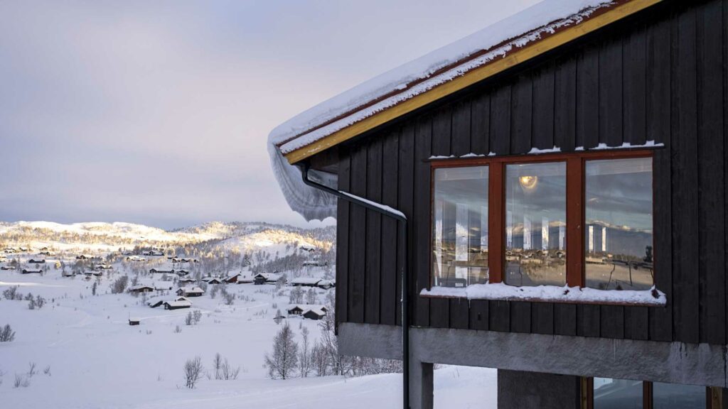 Svart hytte med snødekket tak, utsikt over vinterlandskap og fjell i bakgrunnen, fredelig norsk vinterdag.