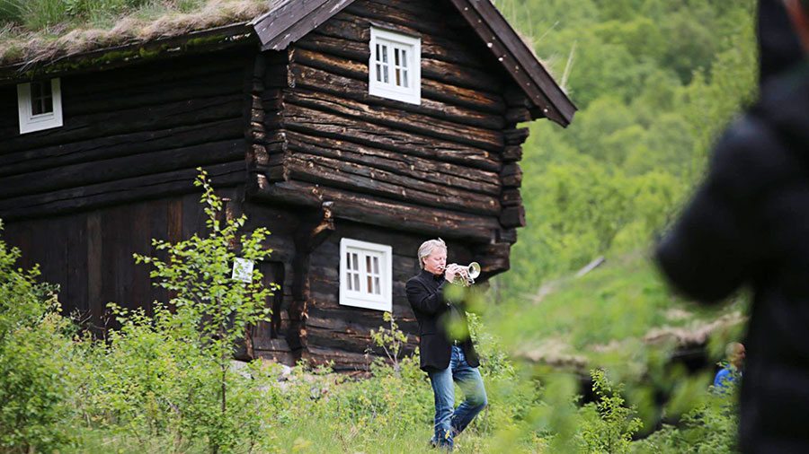 En mann spiller trompet foran en tradisjonell trehytte i et frodig norsk landskap.